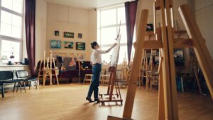 A person painting on a canvas in a sunlit art studio with wooden easels and paintings on the walls.