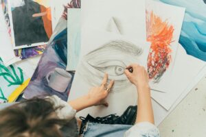 An artist drawing an eye while surrounded by various colorful artworks, using pastels indoors.