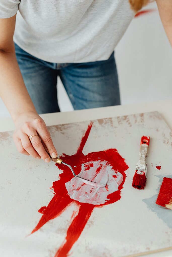 A focused artist uses a palette knife to create red abstract art on canvas.